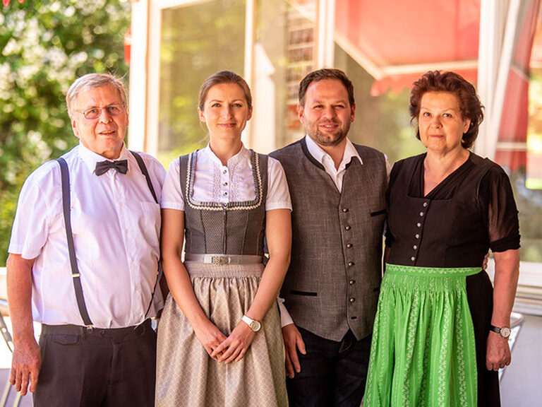Fröhliche Gastgeberfamilie in formeller Kleidung mit Fenstern im Hintergrund auf einer Terasse.